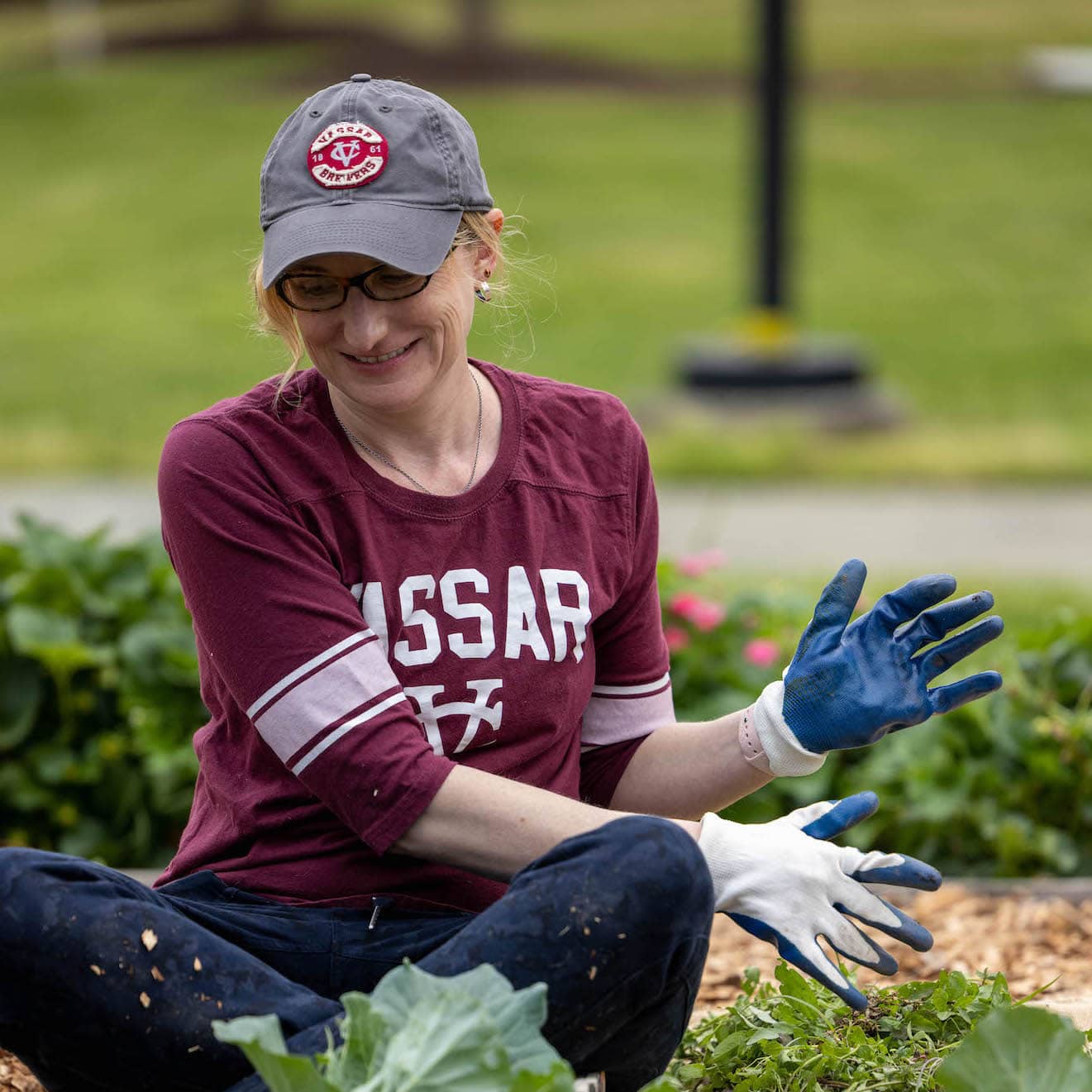 A person in a Vassar jersey and baseball cap, sits on the ground with gloves on, planting flowers.